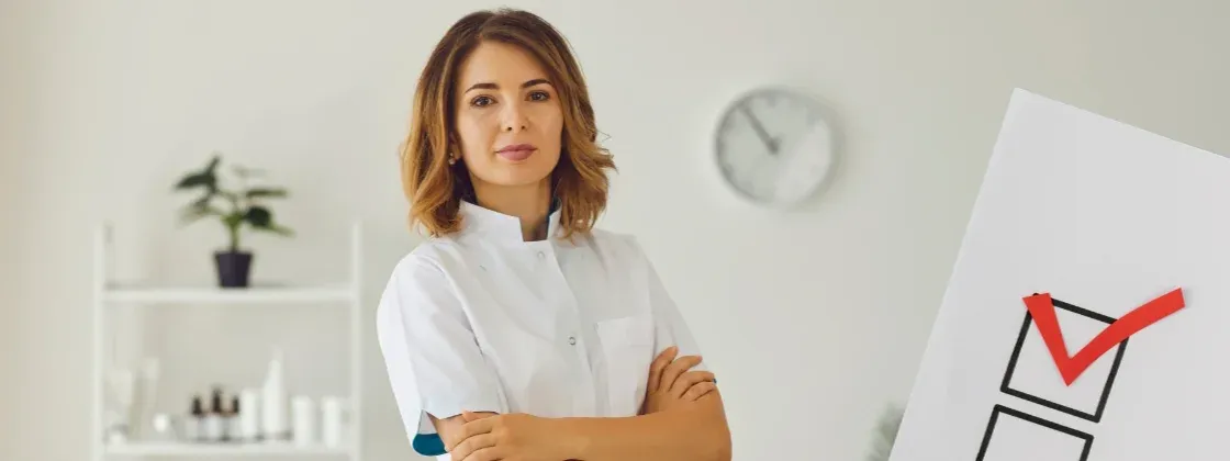 Em primeiro plano, do lado direito, lista de checagem, com um box ticado, faltando dois itens a serem preenchidos. Ao fundo, uma profissional esteticista, com os braços cruzados, olha para a câmera com expressão ansiosa. Cena com cores suaves, variando cinza, bege e o tom de pele branco.